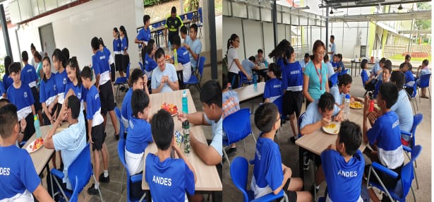 Students in blue Andes uniforms sit and stand around tables eating and socializing in an open cafeteria.